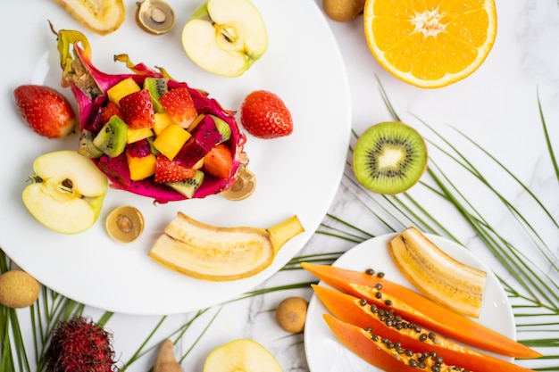 Passievrucht, banaan, longan, tamarinde, appel, papaya op plaat op witte achtergrond met tropische bladeren. Zomer samenstelling, bovenaanzicht. Gezond voedselconcept.