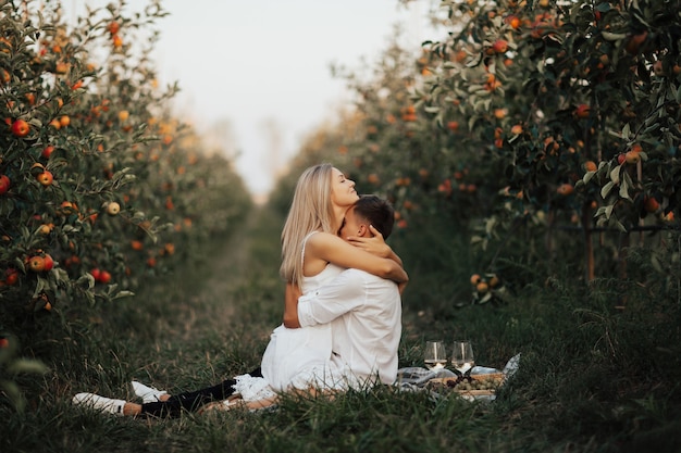 赤いリンゴの果樹園でピクニック毛布に座って抱き締める情熱的な魅力的なカップル。