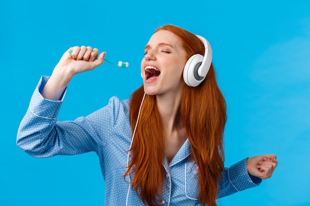 Passionate and carefree cute foxy european redhead woman in pyjama, close eyes wearing headphones, listening music singing into toothbrush, brushing teeth and getting ready morning, blue wall