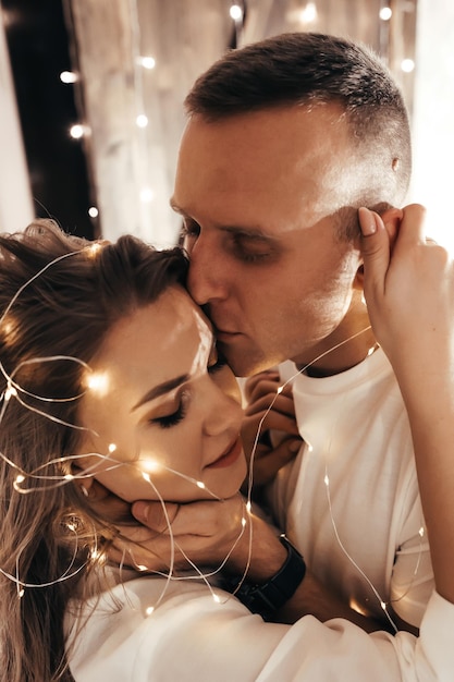 Passionate romantic couple spending time before New Year at home with garlands Young woman and handsome man hug kiss Christmas time