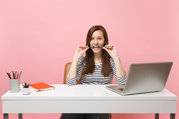 Donna appassionata in abiti casual che rosicchia tenendo la matita tra i denti si siede al lavoro alla scrivania bianca con un computer portatile contemporaneo