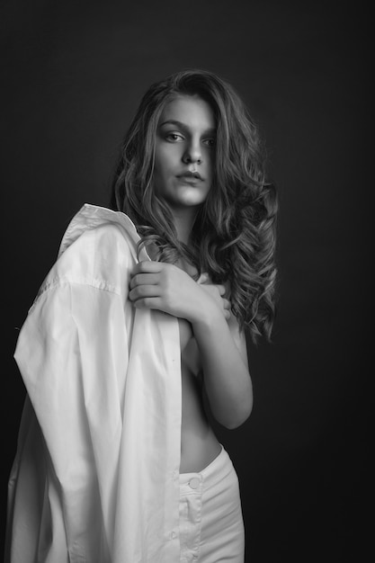 Passionate young model wears white men's shirt and posing with naked shoulders at studio. Monochrome shot