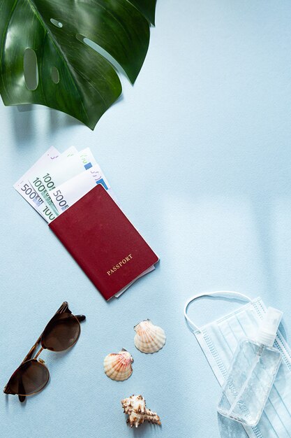 Passport with money, medical face mask and hand sanitizer, sunglasses, seashells and monstera palm leaf.