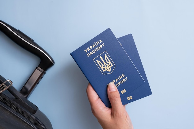 Passports of a citizen of Ukraine in a female hand on a blue background with suitcase closeup Emigration concept Inscription in Ukrainian Ukraine Passport