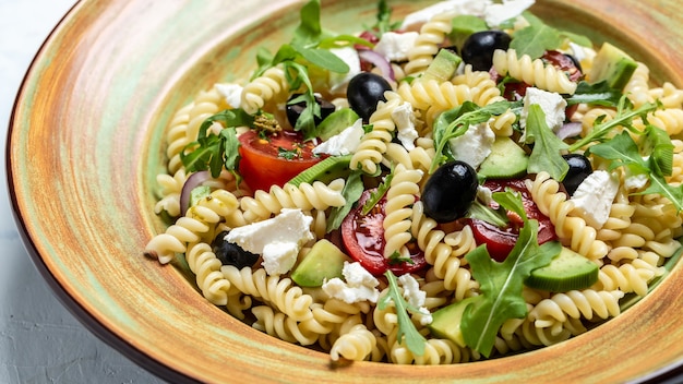 Pasta Greek salad with tomato, avocado, black olives, red onions and cheese feta. Food recipe background. Close up.