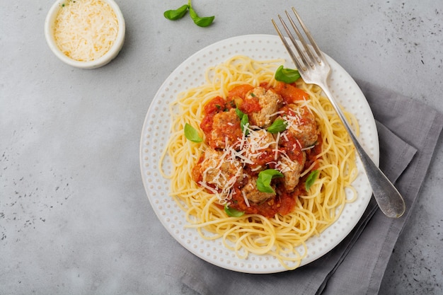 Паста спагетти с томатным соусом, сыром пармезан, базиликом и фрикадельками на белой керамической тарелке на серой бетонной или каменной поверхности. Выборочный фокус.