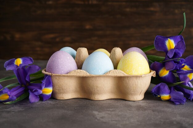 Pastel colorful easter eggs in a cardboard box and fresh flowers on a wooden background.