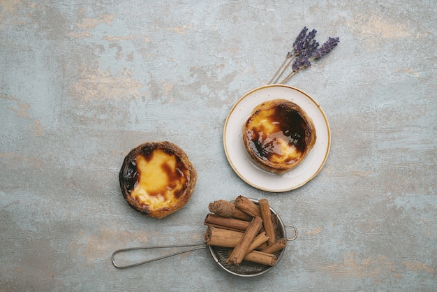 Pastel de nata. Традиционный португальский десерт, яичный пирог на тарелке на деревенском фоне с палочками корицы в ситечке и сушеными веточками лаванды. Вид сверху