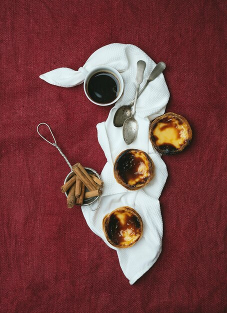 Pastel de nata. Традиционные португальские десертные яичные пироги, чашка кофе и палочки корицы в ситечке на салфетке на текстильном фоне. Вид сверху