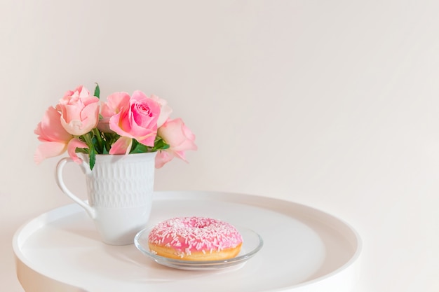 Rose rosa pastello in tazza bianca con la ciambella rosa sul tavolino da salotto bianco