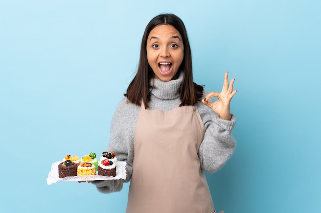 Pastry chef holding a big cake over isolated blue background showing ok sign and thumb up gesture