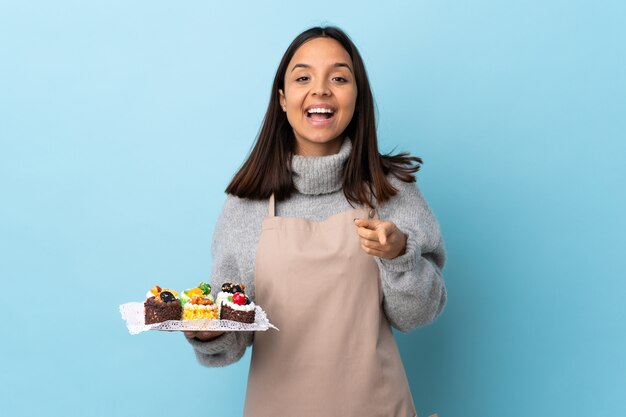 Pastry chef holding a big cake over isolated blue background surprised and pointing front