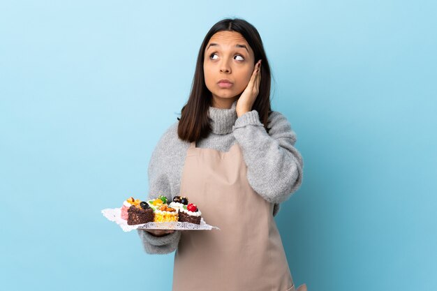 Pastry chef holding a big cake over isolated blue frustrated and covering ears.