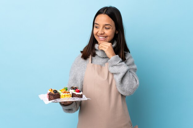 Pastry chef holding a big cake over isolated blue wall looking to the side and smiling