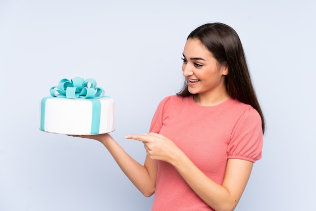 Pastry chef holding a big cake isolated on blue wall pointing to the side to present a product