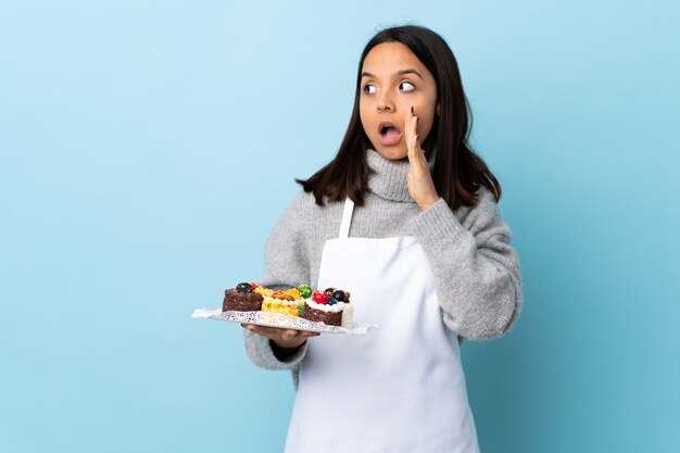 Pastry chef holding a big cake over isolated blue wall whispering something with surprise gesture while looking to the side