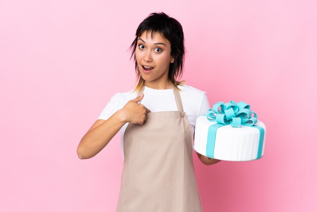 Pastry chef holding a big cake over isolated pink space with surprise facial expression