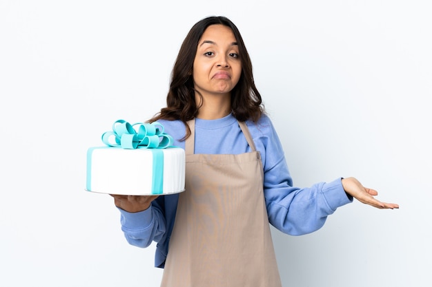 Pastry chef holding a big cake over isolated white background having doubts while raising hands