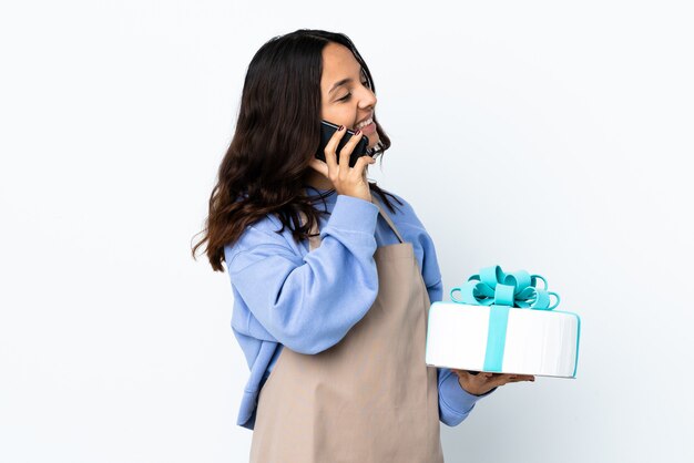 Pastry chef holding a big cake over isolated white wall keeping a conversation with the mobile phone with someone