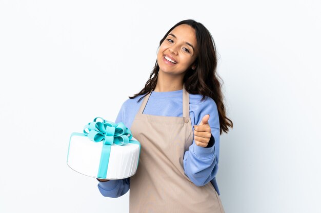 Pastry chef holding a big cake over isolated white wall with thumbs up because something good has happened