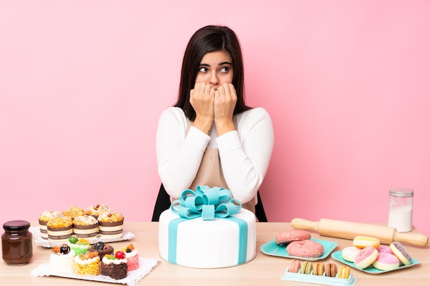 Pastry chef with a big cake in a table over isolated pink wall nervous and scared putting hands to mouth