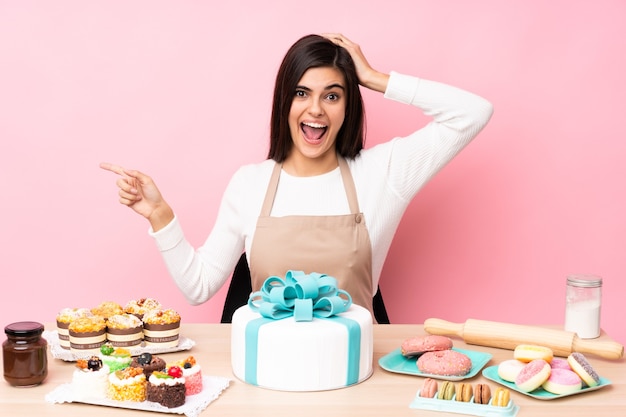 Pastry chef with a big cake in a table over isolated pink wall surprised and pointing finger to the side