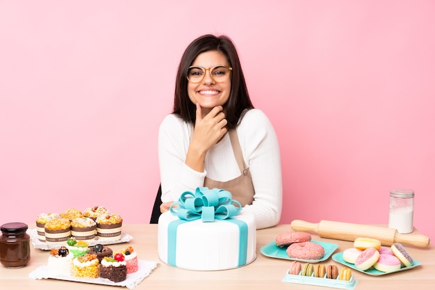 Pastry chef with a big cake in a table over isolated pink wall with glasses and smiling