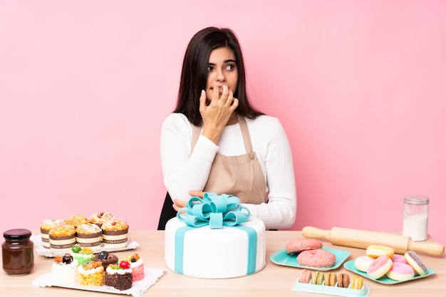 Photo pastry chef with a big cake in a table over pink wall nervous and scared