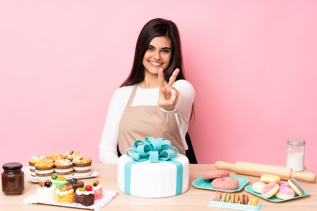 Pastry chef with a big cake in a table over pink wall smiling and showing victory sign