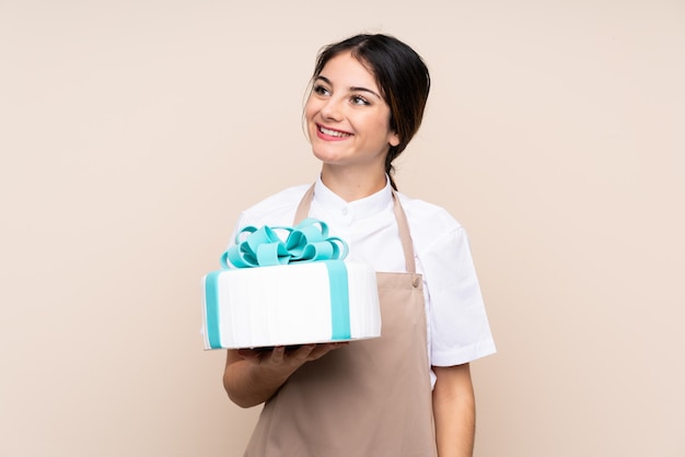 Pastry chef woman holding a big cake over isolated wall looking up while smiling