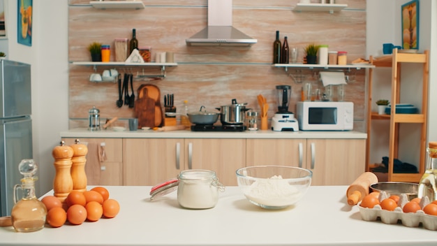 Pastry ingredients ready for cooking in modern kitchen with nobody in it. Modern empty dining room equipped with utensils, fresh eggs and wheat flour in glass bowl for homemade cakes and bread