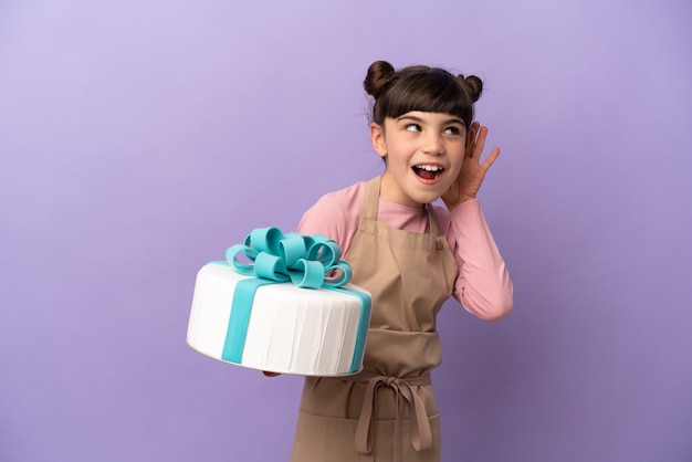 Pastry little girl holding a big cake isolated on purple listening to something by putting hand on the ear