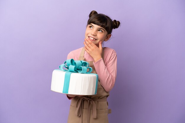 Pasticceria bambina in possesso di una grande torta isolata su viola alzando lo sguardo mentre sorridente