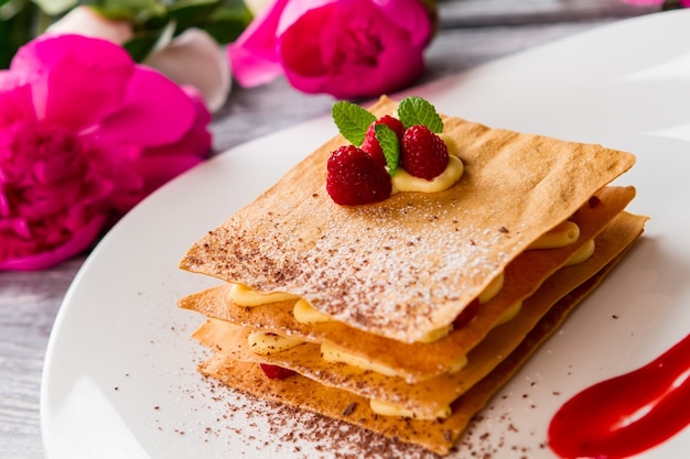 Pastry with raspberries on plate. Pink flowers and mint leaves. Millefeuille with decoration. Crispy dough and juicy berries.