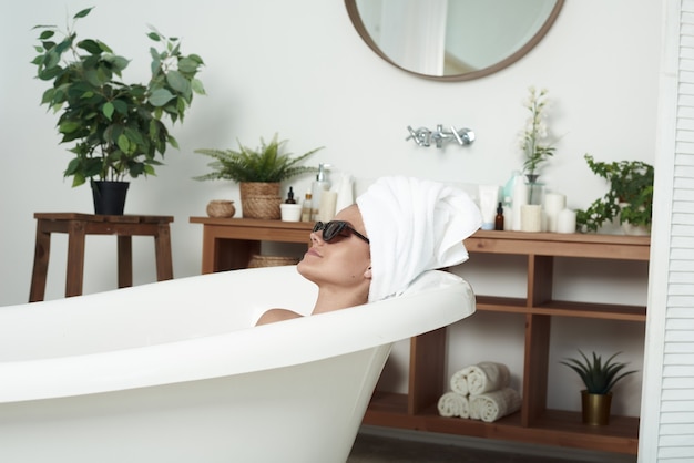 The pathos beautiful girl with vitiligo lies in the bath in the cat's sunglasses and a towel on her head. The concept of fashion, skin care and style.