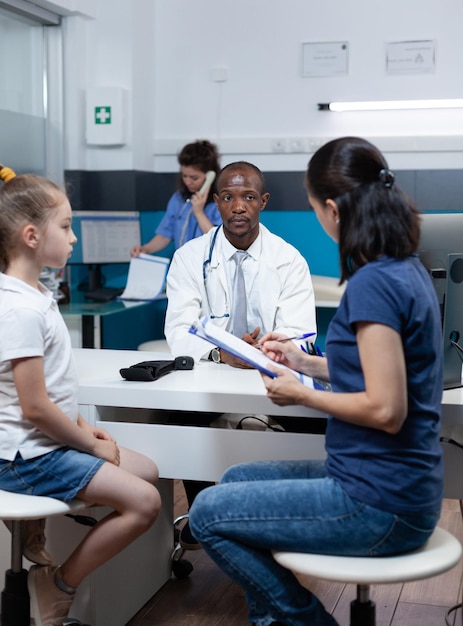 Patient mother writing medical accord on document while discussing sickness expertise with african american therapist doctor during clinical appointment in hospital office. Medicine service