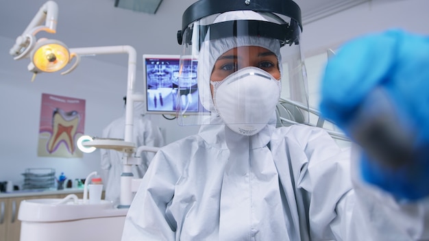 Patient pov of dentist holding dental tools wearing covid protection suit treating patient in new normal clinic. Stomatolog wearing safety gear against coronavirus during heatlhcare check of person.