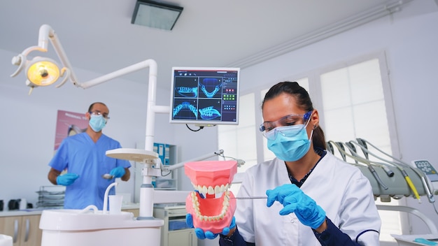 歯科医の患者のハメ撮りは、歯の医療スケルトンアクセサリーを使用して歯科医院で身に着けている歯をきれいにする正しい方法を示しています。ヘルスケアチェック中に保護マスクを着用した歯科医