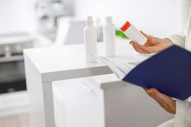 Patient senior blond woman standing near the reception and holding a brochure with beauty treatments.