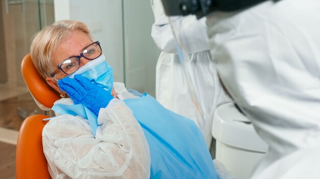 Patient with protection suit touching face having toothache at stomatology clinic during global pandemic. Old woman explaining dental problem to doctor dressed in coverall with face shield and glovel