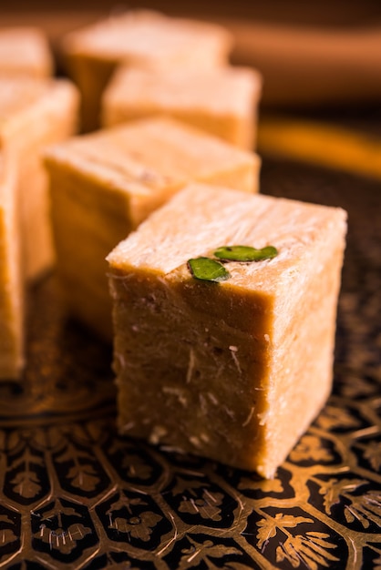 Patisa or Soan Papdi is a popular Indian cube shape flakey and crispy dessert. Served with almonds and pistachio in a plate over moody background. Selective focus