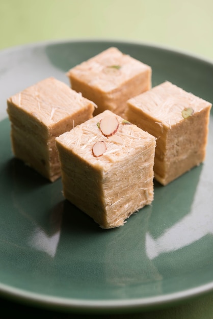 Patisa or Soan Papdi is a popular Indian cube shape flakey and crispy dessert. Served with almonds and pistachio in a plate over moody background. Selective focus