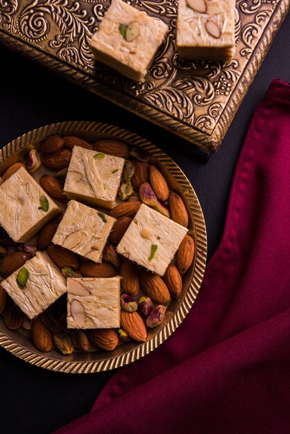 Patisa or Soan Papdi is a popular Indian cube shape flakey and crispy dessert. Served with almonds and pistachio in a plate over moody background. Selective focus