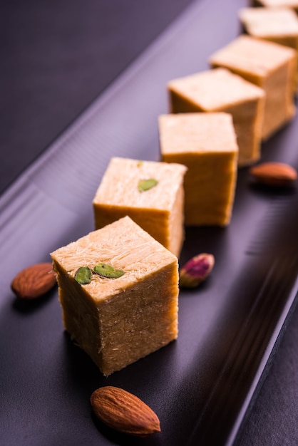 Patisa or Soan Papdi is a popular Indian cube shape flakey and crispy dessert. Served with almonds and pistachio in a plate over moody background. Selective focus