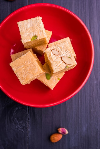 Patisa or Soan Papdi is a popular Indian cube shape flakey and crispy dessert. Served with almonds and pistachio in a plate over moody background. Selective focus