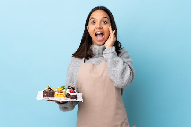 Patissier die een grote cake over geïsoleerd blauw met verrassing en geschokte gezichtsuitdrukking houdt.