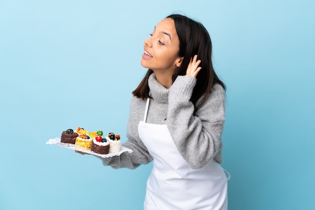 Patissier die een grote cake over geïsoleerde blauwe muur houdt die aan iets luistert door hand op het oor te leggen.