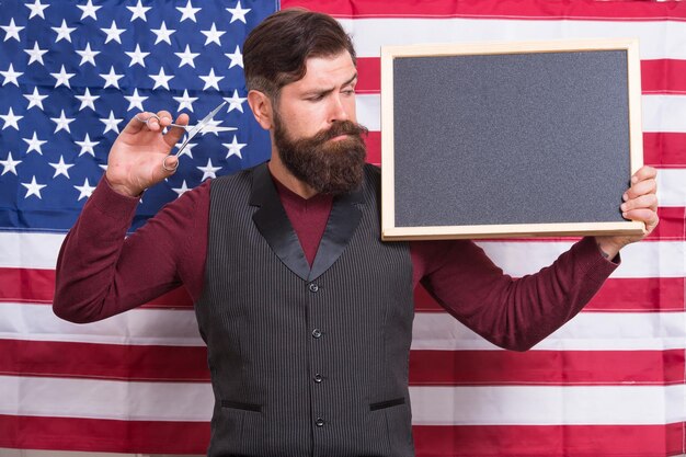 Patriotic education. Bearded man holding scissors and blackboard in education school. Barber education courses. Hipster giving lesson on american flag background. Education and career, copy space.