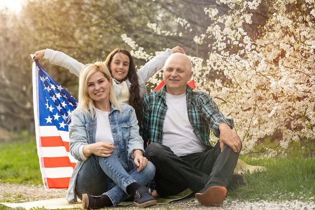 屋外で米国旗を保持している愛国的な家族。緑の背景に米国旗を保持している幸せな高齢白人カップル。