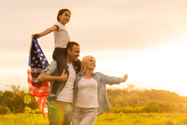 愛国的な休日。幸せな家族、両親と娘の子供たちの女の子が屋外でアメリカ国旗を掲げています。アメリカは7月4日を祝います。